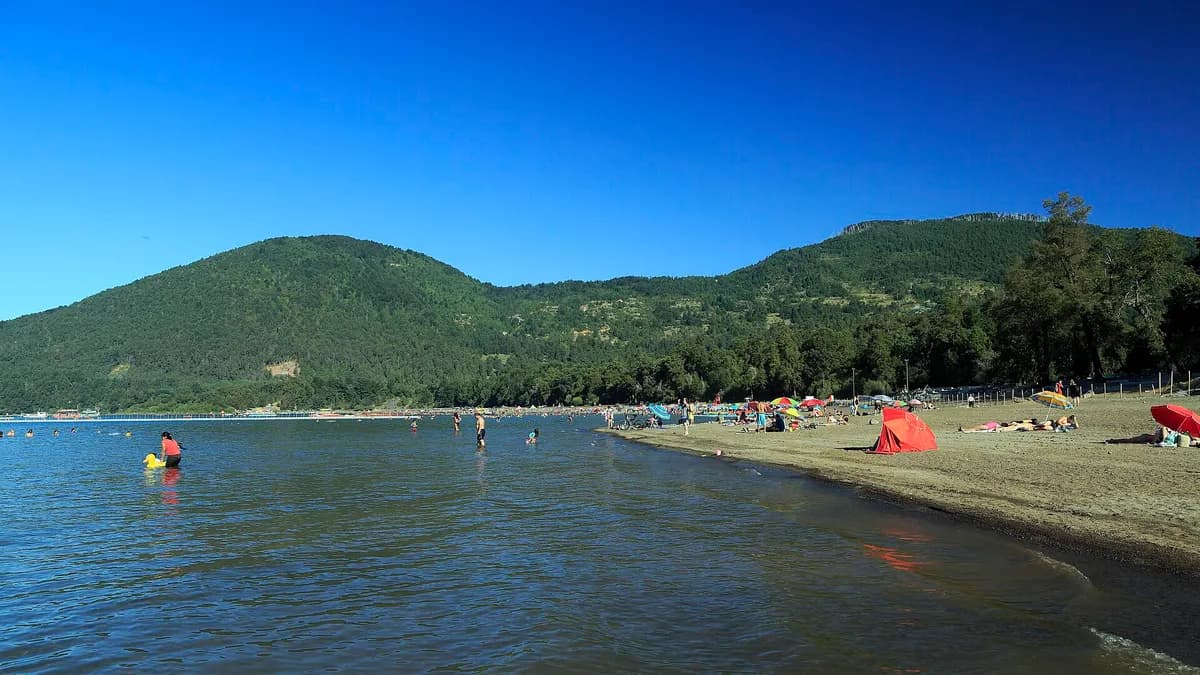 Bañistas y carpas en la Playa Negra del lago Caburgua, con cerros boscosos al fondo
