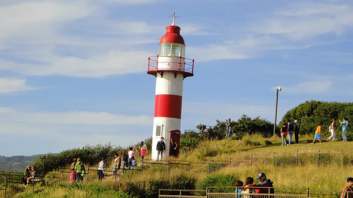Faro rojo y blanco de Niebla sobre una loma verde junto a la costa, con visitantes caminando